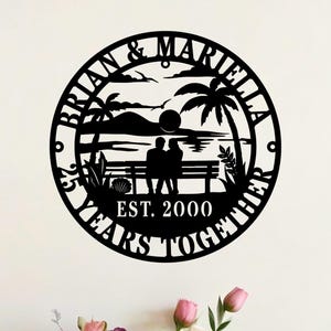 May include: Black metal wall art featuring a circular design with the names "Brian & Mariella" and "55 Years Together". The design includes silhouettes of palm trees, a couple on a bench, and the text "Est. 2000".