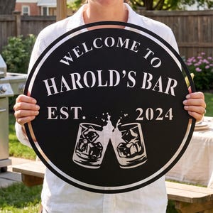Peut inclure: Un panneau circulaire en métal noir avec du texte blanc indiquant WELCOME TO HAROLD'S BAR EST. 2024. Le design comprend un graphique de deux verres qui s'entrechoquent avec des glaçons à l'intérieur, centré dans un fin cadre circulaire.