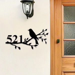 Peut inclure: Panneau d'adresse en métal noir avec le numéro 521, une silhouette d'oiseau perché sur une branche avec des feuilles. Le panneau est fixé sur un mur blanc, à côté d'une lampe extérieure noire et d'une porte en bois.