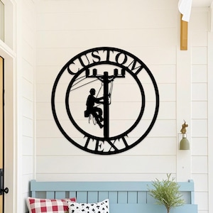 May include: Black metal wall art featuring a silhouette of a lineman working on a utility pole, enclosed in a circular frame. The words "CUSTOM TEXT" are arched around the top and bottom of the circle. The art is mounted on a white wall.