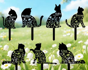 May include: Black metal cat silhouette garden stakes with floral designs. Each stake features a cat's name and dates. The text "Cat Breeds" is in yellow script at the top. The stakes are in a field of white flowers.