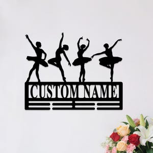 May include: Black metal wall art featuring four ballet dancers in various poses. The dancers are above a rectangular frame with the text "CUSTOM NAME" and three horizontal lines. A decorative piece for a dance studio or bedroom.