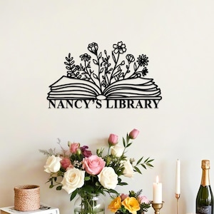 May include: Black metal wall art featuring an open book with floral accents and the text "NANCY'S LIBRARY." The design includes various flower and leaf shapes. The artwork is mounted on a white wall.