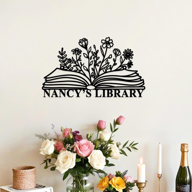 May include: Black metal wall art featuring an open book with floral accents and the text "NANCY'S LIBRARY." The design includes various flower and leaf shapes. The artwork is mounted on a white wall.