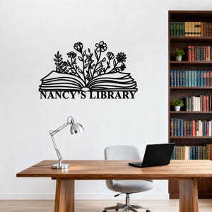 May include: Black metal wall art featuring an open book with floral accents and the text "NANCY'S LIBRARY". The artwork is mounted on a white wall above a wooden desk with a silver lamp and a laptop.