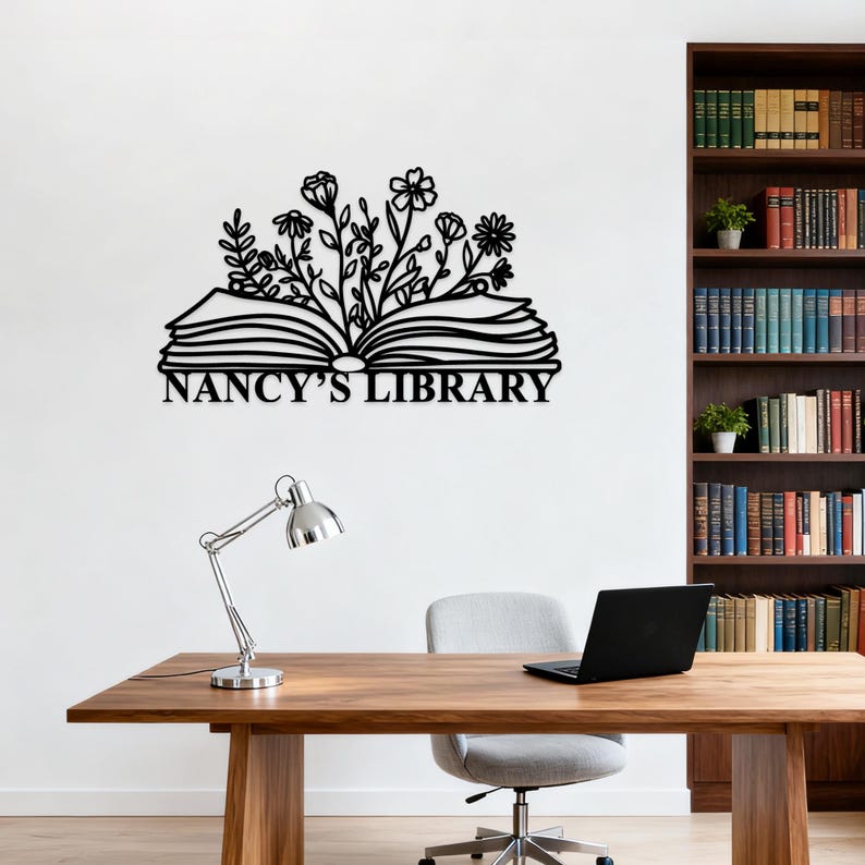 May include: Black metal wall art featuring an open book with floral accents and the text "NANCY'S LIBRARY". The artwork is mounted on a white wall above a wooden desk with a silver lamp and a laptop.