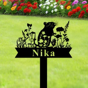 Könnte beinhalten: Schwarzer Metall-Gartenstecker mit einer Ratten-Silhouette und floralen Elementen. Der Name "Nika" ist auf einem Banner am Fuß des Steckers zu sehen. Im Hintergrund ein grüner Rasen und ein Blumenbeet mit bunten Blüten.