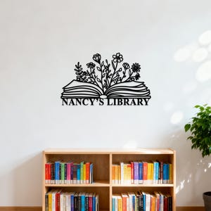 May include: Black metal wall art featuring an open book with floral accents and the text "NANCY'S LIBRARY". The artwork is mounted on a white wall above a wooden bookshelf filled with colorful books.