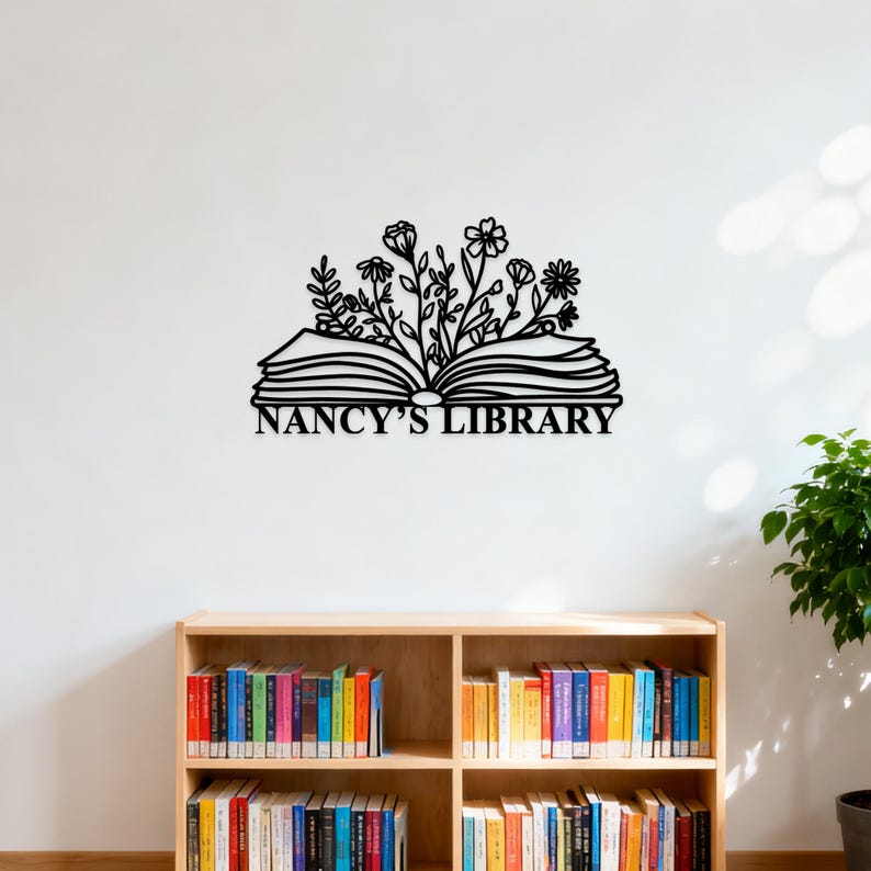 May include: Black metal wall art featuring an open book with floral accents and the text "NANCY'S LIBRARY". The artwork is mounted on a white wall above a wooden bookshelf filled with colorful books.
