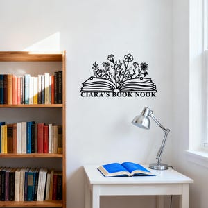 May include: Black metal wall art featuring an open book with floral accents and the text "CIARA'S BOOK NOOK." The artwork is mounted on a white wall, with a wooden bookshelf and a desk with a lamp and an open book in the background.
