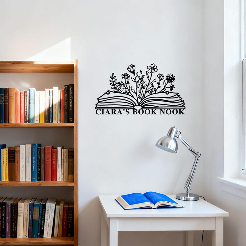 May include: Black metal wall art featuring an open book with floral accents and the text "CIARA'S BOOK NOOK." The artwork is mounted on a white wall, with a wooden bookshelf and a desk with a lamp and an open book in the background.
