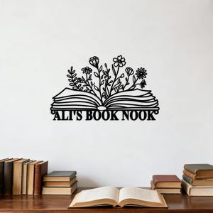 May include: Black metal wall art featuring an open book with floral accents. The text "ALI'S BOOK NOOK" is displayed below the book. The image includes a wooden table with several books.