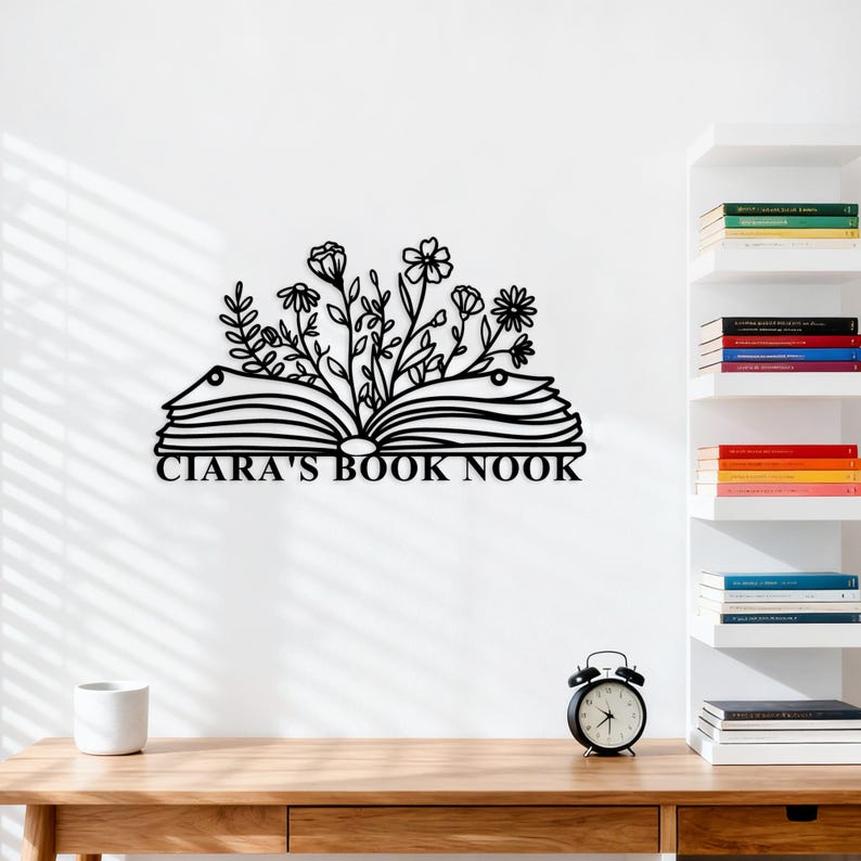 May include: Black metal wall art depicting an open book with floral accents. The text "CIARA'S BOOK NOOK" is centered below the book. The art is displayed on a white wall above a wooden desk, with a white mug and a black alarm clock.