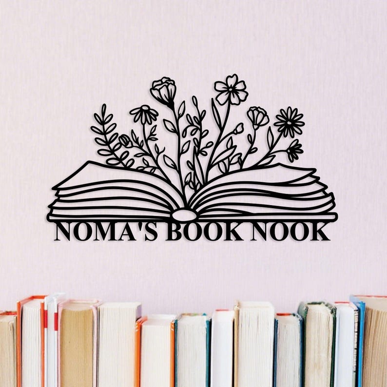 May include: Black metal wall art featuring an open book with floral accents and the text "NOMA'S BOOK NOOK". The artwork is set against a light pink background, with a row of books at the bottom.