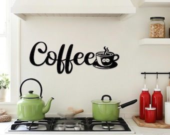 May include: Black metal wall art spelling "Coffee" with a coffee cup design. The sign is mounted on a white wall above a stove with green pots and a kettle. Kitchen decor.