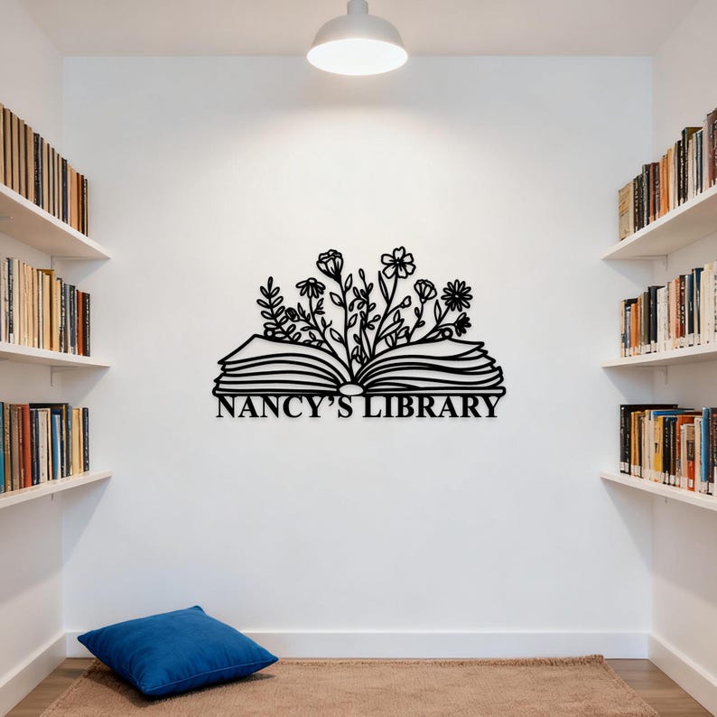 May include: Black metal wall art featuring an open book with floral accents and the text "NANCY'S LIBRARY". The artwork is mounted on a white wall in a room with bookshelves and a blue cushion.