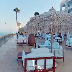 May include: Outdoor seating area with white sofas, wooden tables, and macrame umbrellas. A tall palm tree and a lighthouse are visible in the background. The text "THE SEVEN WOMEN ARTISTRY IN HOME DESIGN" is at the bottom.