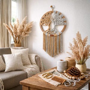 Op de afbeelding: Een macramé wandkleed met een boomontwerp en een houten frame. De boom is gemaakt met beige en grijs garen, met franjes eronder. De decoratie wordt gepresenteerd in een gezellige woonkamer met neutrale tinten en natuurlijke elementen.