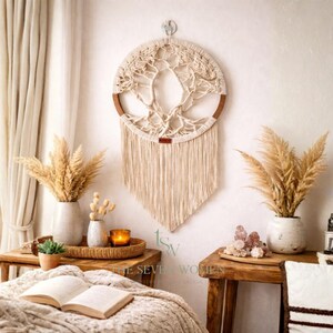 Op de afbeelding: Een beige macramé wandkleed met een levensboomontwerp en een houten ring. De wanddecoratie wordt getoond in een kamer met pampasgras in vazen en een brandende kaars.