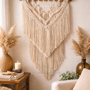 Op de afbeelding: Crèmekleurige macramé wandhanger met een geometrisch ontwerp en franjes. Het textielkunstwerk is opgehangen aan een houten staaf. De wanddecoratie wordt getoond in een kamer met neutrale tinten, een kaars en decoratieve vazen.