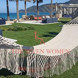 May include: A cream-coloured hammock with a fringed edge, suspended between two wooden supports. The hammock is set on a green lawn with a view of the ocean and palm trees in the background. The text "SEVEN WOMEN" is visible on the hammock.