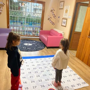 May include: Two children stand on a large white and red number mat with a blue circle in the center. The mat has numbers from 1 to 100 in rows. The children are wearing casual clothing. The mat is on a wooden floor.