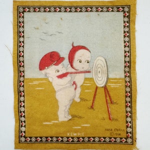 May include: A vintage fabric patch featuring two Kewpie dolls in red and white clothing, aiming at a target with a bow and arrow. The patch has a gold background with a brown and white border and the text "Kewpie" and "Rose O'Neill 1914".