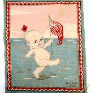 May include: A vintage fabric patch featuring a cartoon baby holding an American flag. The baby is wearing a red hat and is standing in the water. The patch has a blue background with a pink sky and a red, white, and blue border. The text "Rose Snell 1944" is printed in the bottom right corner.