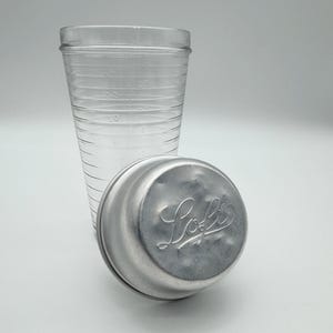 Op de afbeelding: Een heldere glazen maatbeker met afgestudeerde markeringen en een zilveren metalen deksel. Op het deksel staat het woord "Loft" in een cursief lettertype. De beker is ontworpen voor het meten van vloeistoffen en het deksel is waarschijnlijk voor opslag.