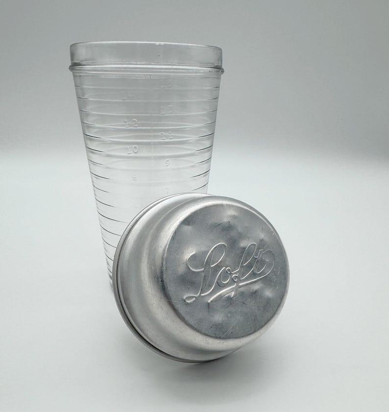 Op de afbeelding: Een heldere glazen maatbeker met afgestudeerde markeringen en een zilveren metalen deksel. Op het deksel staat het woord "Loft" in een cursief lettertype. De beker is ontworpen voor het meten van vloeistoffen en het deksel is waarschijnlijk voor opslag.