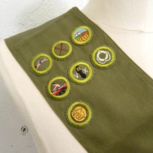 May include: Olive green sash with embroidered patches. Patches feature various designs, including a swimmer, a mountain landscape, and a life preserver. The sash is displayed on a mannequin.