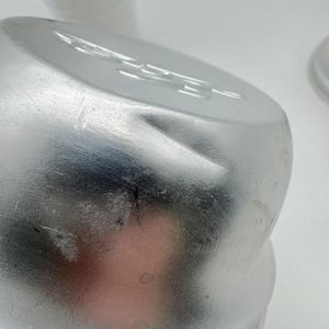 Op de afbeelding: Close-up van een zilveren metalen container met een licht gedeukt oppervlak. De container heeft een afgeronde bovenkant en een geribbelde basis. De achtergrond bevat een stapel witte papieren bekers.
