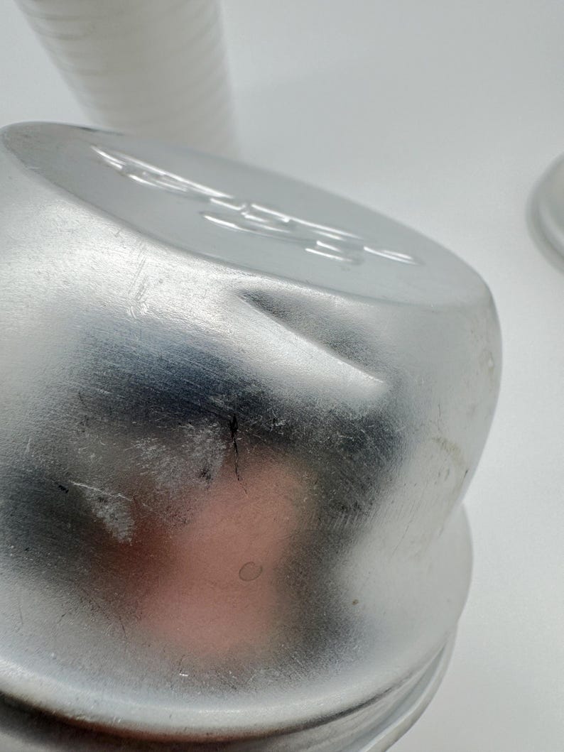 Op de afbeelding: Close-up van een zilveren metalen container met een licht gedeukt oppervlak. De container heeft een afgeronde bovenkant en een geribbelde basis. De achtergrond bevat een stapel witte papieren bekers.