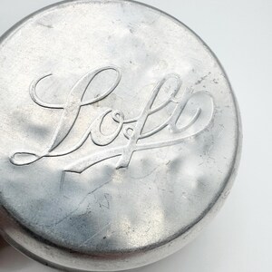 Op de afbeelding: Close-up van een ronde, zilverkleurige metalen deksel. Het woord "Loft" is in een cursief lettertype in het midden in reli&euml;f aangebracht. De deksel heeft een licht getextureerd oppervlak en een afgeronde rand, tegen een witte achtergrond.