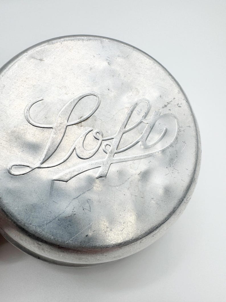 Op de afbeelding: Close-up van een ronde, zilverkleurige metalen deksel. Het woord "Loft" is in een cursief lettertype in het midden in reli&euml;f aangebracht. De deksel heeft een licht getextureerd oppervlak en een afgeronde rand, tegen een witte achtergrond.