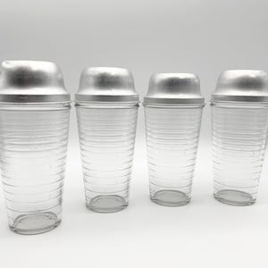 Op de afbeelding: Vier vintage cocktailshakers met heldere glazen bodies en zilverkleurige metalen deksels. De shakers zijn van verschillende formaten, met horizontale geribbelde details op het glas. De grootste shaker heeft maatstreepjes.