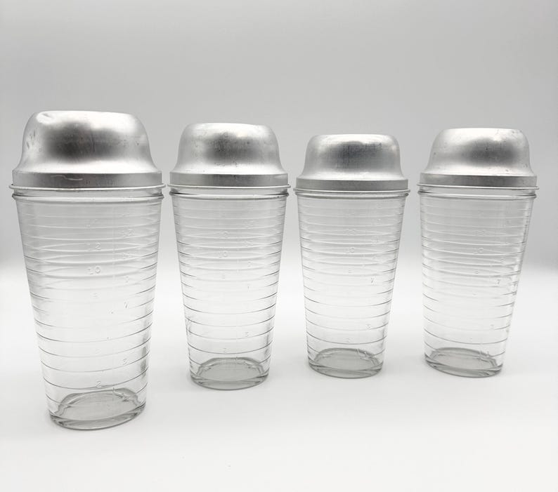 Op de afbeelding: Vier vintage cocktailshakers met heldere glazen bodies en zilverkleurige metalen deksels. De shakers zijn van verschillende formaten, met horizontale geribbelde details op het glas. De grootste shaker heeft maatstreepjes.