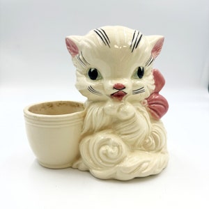 May include: A white ceramic cat planter with a pink bow and a separate cream-colored pot. The cat has black eyes and a black nose.