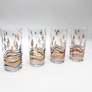 Puede incluir: Juego de cuatro vasos transparentes con un diseño decorativo. Los vasos presentan un patrón de peces dorados y negros, con bandas doradas en forma de olas en la base. Los vasos son altos y cilíndricos.