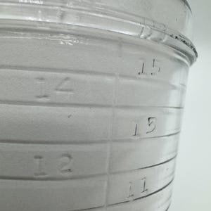 Op de afbeelding: Close-up van een doorzichtige glazen maatbeker met in reli&euml;f aangebrachte horizontale lijnen en numerieke markeringen. De cijfers 10, 11, 12, 14 en 15 zijn zichtbaar. De beker heeft een brede, cilindrische vorm.