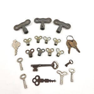 May include: A selection of antique keys and clockwork keys in different shapes and sizes. The metal keys show signs of wear and tear, with designs including T-shapes, circular, and decorative styles. The keys are arranged on a white background.