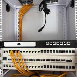 Könnte beinhalten: Ein offenes Server-Rack mit Netzwerkausrüstung. Das Rack enthält mehrere Netzwerk-Switches, ein weißes Gerät und gelbe Netzwerkkabel. Zwei schwarze Lüfter sind oben montiert, und ein schwarzes Netzkabel ist sichtbar.