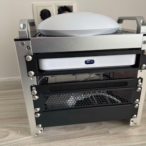 Könnte beinhalten: Ein silbernes Metallregal mit drei Regalböden. Im oberen Regal befindet sich ein weißer WLAN-Router. Im mittleren Regal befindet sich eine weiße Box mit einem blauen Etikett. Das untere Regal ist schwarz mit einem Gitterdesign.