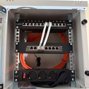 Könnte beinhalten: Eine weiße Metallbox mit einer schwarzen Steckdosenleiste, zwei Netzwerkschaltern und orangefarbenen und weißen Ethernet-Kabeln. Die Box ist mit "DIGITUS" beschriftet.