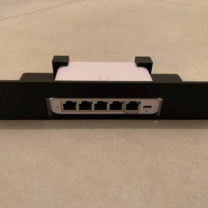 Könnte beinhalten: Schwarzer Metall-Rack-Halterung mit einem weißen Netzwerkgerät. Das Netzwerkgerät hat fünf Ethernet-Anschlüsse und einen USB-C-Anschluss. Die Rack-Halterung hat Befestigungslöcher an jedem Ende.