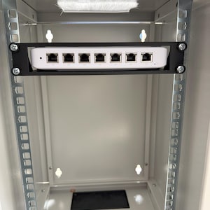 Könnte beinhalten: Ein weißes Netzwerkgerät mit mehreren Anschlüssen ist in einem Metallrack montiert. Das Gerät wird von einer schwarzen Halterung gehalten. Das Rack befindet sich in einem weißen Schrank mit einem schwarzen rechteckigen Lüfter unten.