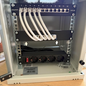 Könnte beinhalten: Ein weißer Metall-Netzwerk-Rack mit einem schwarzen 12-Port CAT6A Ethernet-Patchpanel, einer schwarzen Steckdosenleiste mit vier Steckdosen und weißen Ethernet-Kabeln. Der Rack hat ein Etikett mit der Aufschrift "deleyCON".