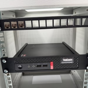 Könnte beinhalten: Ein schwarzer ThinkCentre-Computer ist in einem Serverrack montiert. Die Vorderseite des Computers hat Anschlüsse und das ThinkCentre-Logo. Oberhalb des Computers befindet sich eine schwarze Blende mit Netzwerkanschlüssen.