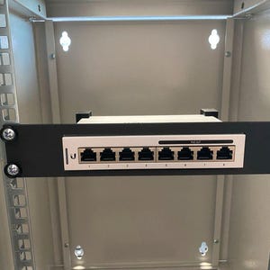 Könnte beinhalten: Ein schwarzer Metallrack mit einem weißen 8-Port-Netzwerkschalter, der darin montiert ist. Der Schalter trägt ein Etikett mit der Aufschrift "POE-8GT".