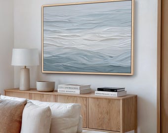 May include: A framed abstract painting depicting ocean waves in shades of blue and white. The artwork has a textured appearance, with visible brushstrokes creating a sense of depth. The frame is a light wood color, complementing the cool tones of the painting.